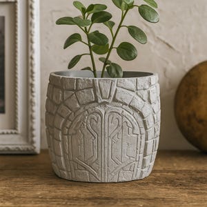 May include: A gray, stone-textured planter with a decorative arched design, holding a green leafy plant. The planter has a cylindrical shape and sits on a wooden surface. The design resembles a stone wall.