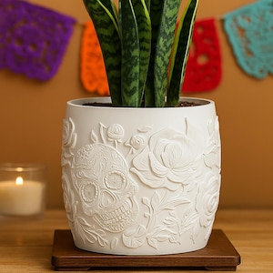 May include: A white ceramic planter with a detailed sugar skull and floral relief design. The planter holds a green snake plant and sits on a wooden base. A lit candle and colorful papel picado banners are in the background.