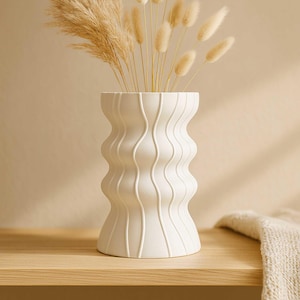 Pode incluir: Um vaso de cerâmica branco com um design ondulado e texturizado. O vaso contém ervas ornamentais secas e caules fofos de cor clara. O vaso está sobre uma superfície de madeira, criando uma estética neutra e natural.