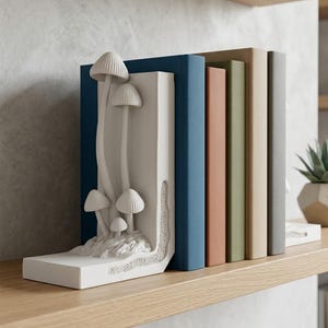 May include: A white mushroom-shaped bookend featuring detailed fungi sculptures. It supports a row of books with blue, peach, green, beige, and grey spines. The items are displayed on a light wood shelf.