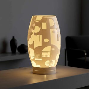 May include: A cream-colored, oval-shaped table lamp with a geometric pattern. The lamp is illuminated, showcasing a design of shapes and lines. The lamp sits on a matching base and is plugged in. The background includes a dark shelf with black vases.