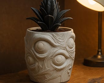 May include: A unique, off-white ceramic planter with a detailed, textured design of multiple eyes. The planter holds a dark green, spiky succulent. The planter is sitting on a wooden surface, with a lamp in the background.