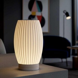 May include: A modern, off-white table lamp with a vertically striped design. The lamp has a cylindrical shape with a rounded top and base. The lamp is lit, emitting a soft glow. The base is a light gray color. The lamp is on a gray surface.