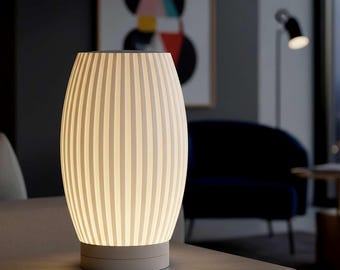 Puede incluir: Lámpara de mesa moderna de color blanco roto con un diseño de rayas verticales. La lámpara tiene forma cilíndrica con la parte superior e inferior redondeadas. Emite una luz suave. La base es de color gris claro. La lámpara está sobre una superficie gris.