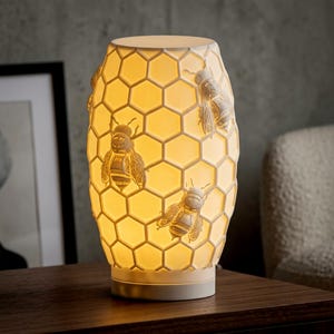 May include: A cylindrical table lamp featuring a white, translucent honeycomb pattern exterior with embossed bee figures. The lamp emits a warm, soft glow from within, highlighting the hexagonal structure and the detailed bee motifs on its surface.