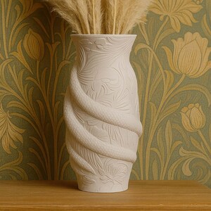 May include: A cream-coloured ceramic vase with a snake design, adorned with detailed floral and leaf patterns. The vase holds dried pampas grass. The background features a green and gold floral wallpaper.