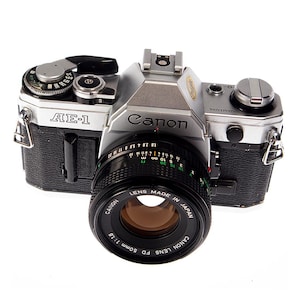 May include: A silver and black Canon AE-1 camera with a 50mm lens. The lens is black with white text, and the camera body has the Canon logo. The camera has various dials and settings.