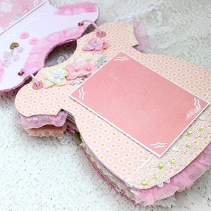 May include: A pink and yellow baby dress scrapbook with a pink card with a white border and a white wavy line design. The scrapbook is decorated with lace, flowers, and polka dots.