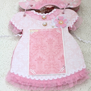 May include: Pink fabric baby dress with white lace trim and a pink and white floral embellishment. The dress has a blank pink card inside for a photo or message.