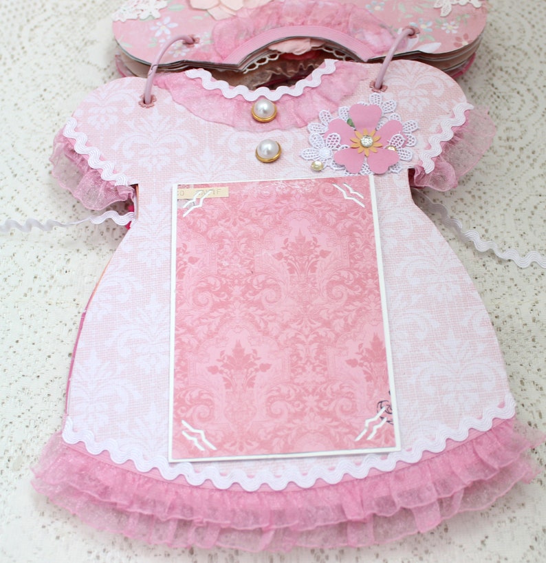 May include: Pink fabric baby dress with white lace trim and a pink and white floral embellishment. The dress has a blank pink card inside for a photo or message.