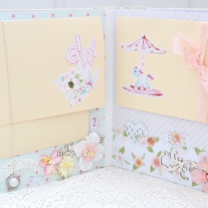 May include: A pink and white scrapbook with three pages. The first page features a pink "Girl" sticker and a white star sticker. The second page is blank. The third page has a sticker of a carousel with a pink and white striped canopy and a white horse. The pages are decorated with lace, flowers, and ribbons.