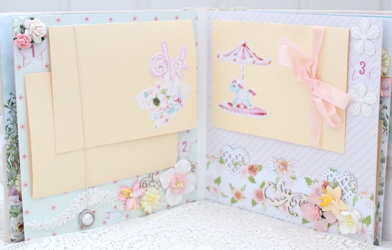 May include: A pink and white scrapbook with three pages. The first page features a pink "Girl" sticker and a white star sticker. The second page is blank. The third page has a sticker of a carousel with a pink and white striped canopy and a white horse. The pages are decorated with lace, flowers, and ribbons.