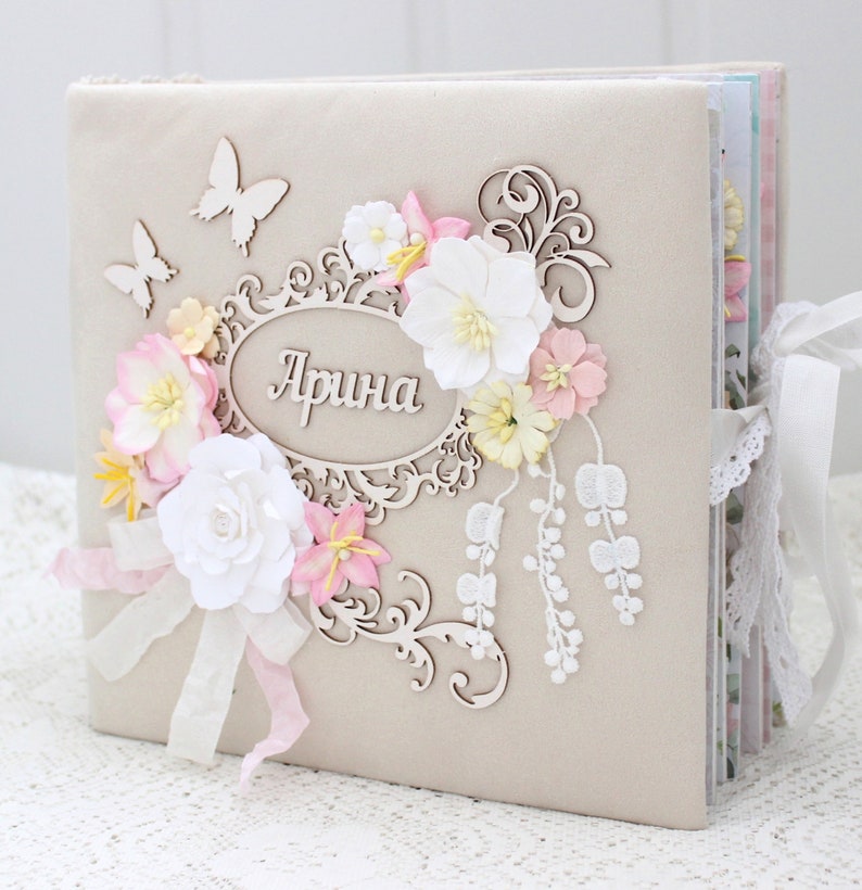May include: A beige scrapbook with a decorative wooden frame and floral embellishments. The frame has the name "Арина" written in cursive. The scrapbook is tied with a white lace ribbon.