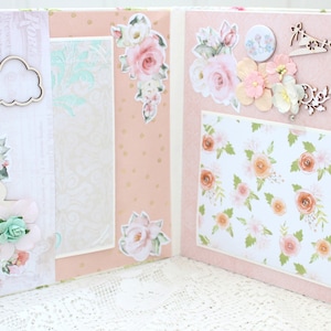 May include: A pink scrapbook with floral decorations, including a silhouette of a pregnant woman, clouds, and a floral pattern. The scrapbook is open to reveal a page with a floral pattern and a wooden cutout of a dress hanging on a clothesline.