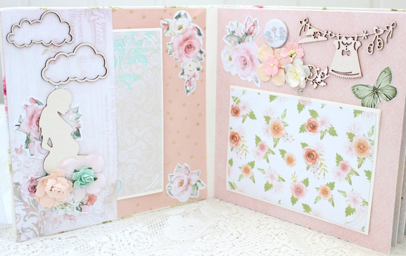May include: A pink scrapbook with floral decorations, including a silhouette of a pregnant woman, clouds, and a floral pattern. The scrapbook is open to reveal a page with a floral pattern and a wooden cutout of a dress hanging on a clothesline.
