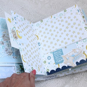 May include: A baby scrapbook with a blue and white color scheme. The scrapbook features a variety of paper textures and patterns, including stars, moons, and clouds. The scrapbook is open to a page with a pocket and a small crib with a baby inside. The text on the page reads "Sleep so soundly every night, you are my little cuddlebug".