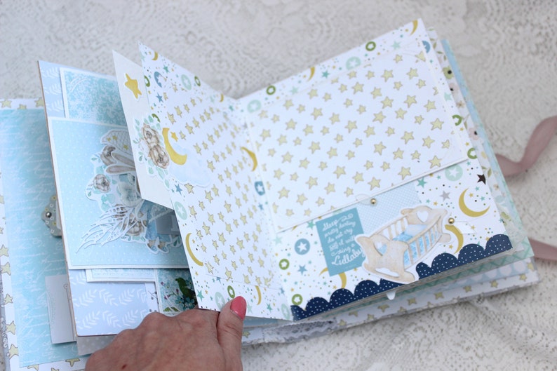 May include: A baby scrapbook with a blue and white color scheme. The scrapbook features a variety of paper textures and patterns, including stars, moons, and clouds. The scrapbook is open to a page with a pocket and a small crib with a baby inside. The text on the page reads "Sleep so soundly every night, you are my little cuddlebug".