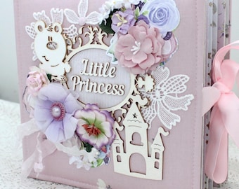 May include: A pink scrapbook with a white lace design and a wooden cutout of a castle. The scrapbook has a pink ribbon tied around it and the words "Little Princess" are written on the front.