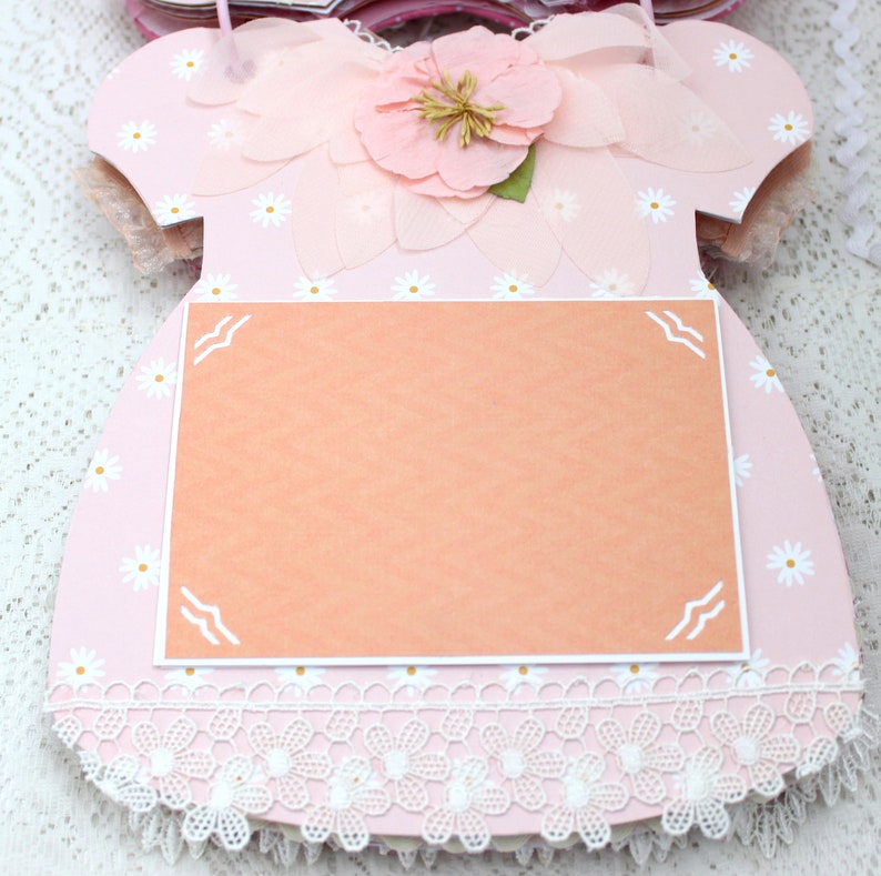 May include: A pink paper dress with a white lace trim and a blank peach-colored card in the center. The dress is decorated with white daisies and a pink flower with a yellow center.