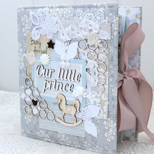 May include: A gray fabric scrapbook with a pink ribbon. The cover is decorated with white lace, flowers, and a wooden rocking horse. The text "Our little prince" is written on the cover.