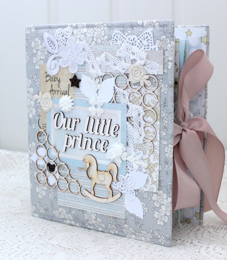 May include: A gray fabric scrapbook with a pink ribbon. The cover is decorated with white lace, flowers, and a wooden rocking horse. The text "Our little prince" is written on the cover.