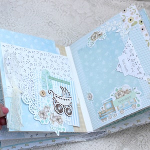 May include: A blue and white scrapbook with decorative paper and embellishments. The scrapbook has pages with the text "first tooth" and "first steps".