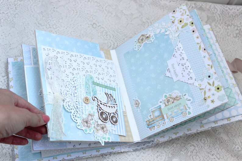 May include: A blue and white scrapbook with decorative paper and embellishments. The scrapbook has pages with the text "first tooth" and "first steps".