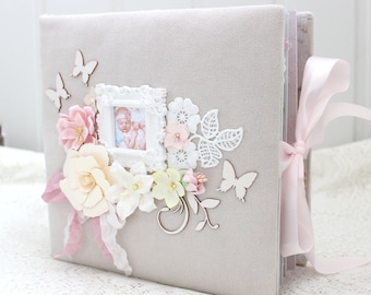 May include: A gray fabric scrapbook with a pink ribbon, white lace, and paper flowers. A white frame with a photo of a baby girl is in the center of the cover. The scrapbook is decorated with white butterflies.
