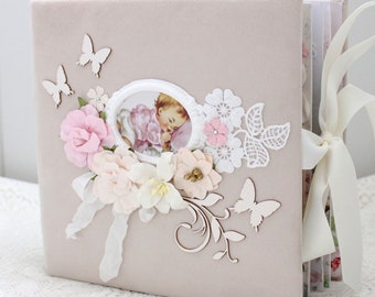 May include: A pink velvet scrapbook with a white lace design, a picture of a baby, and a white ribbon. The scrapbook is decorated with flowers, butterflies, and a wooden swirl design.