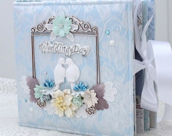 May include: A light blue wedding album with a white satin ribbon. The cover features a decorative frame with the words "Wedding Day" and a pair of white doves. The album is decorated with white and blue flowers.
