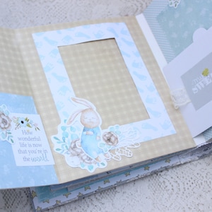 May include: A light blue scrapbook with a beige checkered pattern. The scrapbook has a photo frame with a blue bunny illustration and a tag that says "How wonderful life is now that you're in the world".
