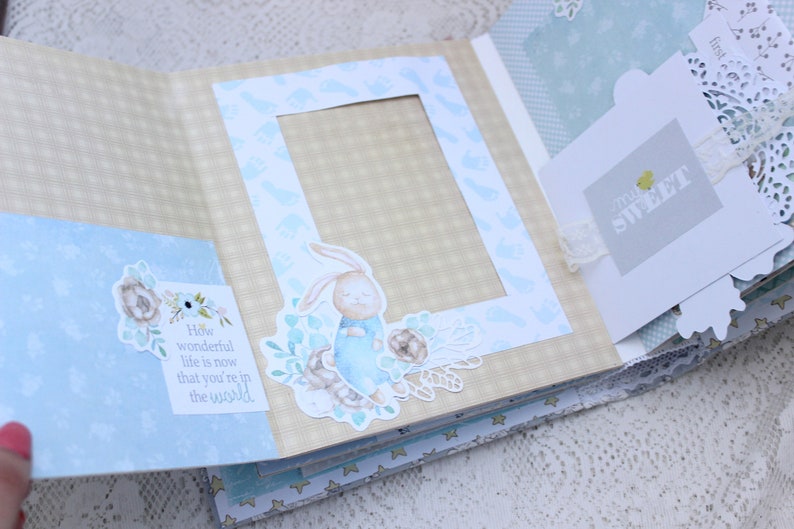 May include: A light blue scrapbook with a beige checkered pattern. The scrapbook has a photo frame with a blue bunny illustration and a tag that says "How wonderful life is now that you're in the world".