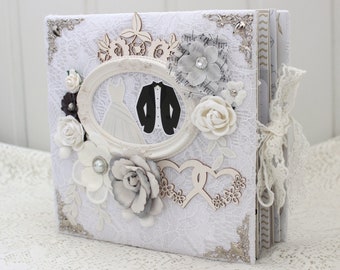 May include: White wedding album with lace trim, a decorative frame with a bride and groom silhouette, and wooden hearts. The album has a white background with a lace overlay and a decorative silver trim.