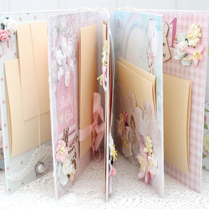 May include: A pink and white scrapbook with pages decorated with flowers, ribbons, and lace. The pages are blank and ready for photos and journaling. The scrapbook has a "Baby Girl" tag on the front.