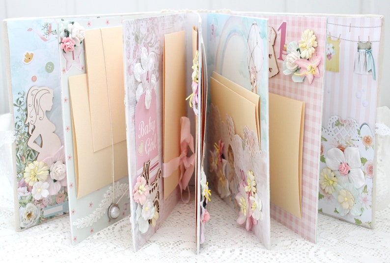 May include: A pink and white scrapbook with pages decorated with flowers, ribbons, and lace. The pages are blank and ready for photos and journaling. The scrapbook has a "Baby Girl" tag on the front.