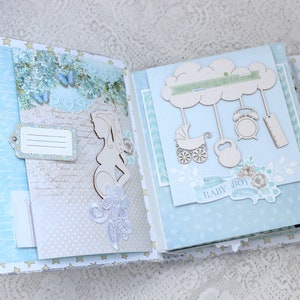May include: A blue and white scrapbook with a wooden cutout of a pregnant woman on the left page and a wooden cutout of a cloud with hanging baby toys on the right page. The scrapbook has a light blue background with white polka dots and a white ribbon.