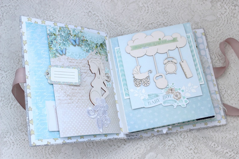 May include: A blue and white scrapbook with a wooden cutout of a pregnant woman on the left page and a wooden cutout of a cloud with hanging baby toys on the right page. The scrapbook has a light blue background with white polka dots and a white ribbon.