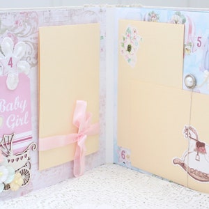 May include: A pink and white scrapbook with pages for photos and journaling. The pages are decorated with flowers, ribbons, and other embellishments. The scrapbook has the words "Baby Girl" on the front.