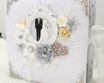 May include: White wedding album with lace overlay, decorated with flowers, a heart, and a framed illustration of a bride and groom.