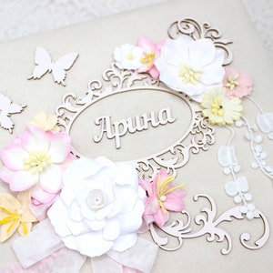 May include: A white scrapbook with a decorative wooden frame and the name "Арина" in cursive. The frame is adorned with white and pink paper flowers, butterflies, and lace.