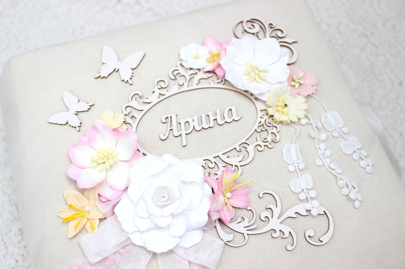 May include: A white scrapbook with a decorative wooden frame and the name "Арина" in cursive. The frame is adorned with white and pink paper flowers, butterflies, and lace.