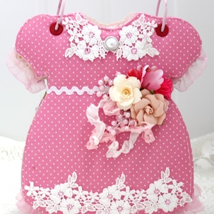 May include: A pink fabric baby dress with white polka dots and lace trim. The dress has a pearl button and a flower embellishment.