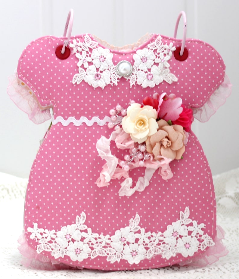 May include: A pink fabric baby dress with white polka dots and lace trim. The dress has a pearl button and a flower embellishment.
