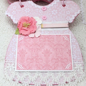 May include: Pink baby dress scrapbook with a lace trim, a pink flower embellishment, and a white card with the text "Sweet Baby Girl".