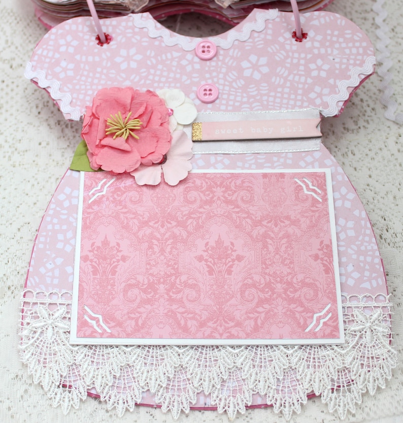 May include: Pink baby dress scrapbook with a lace trim, a pink flower embellishment, and a white card with the text "Sweet Baby Girl".