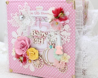 May include: Pink polka-dot scrapbook album with lace trim and gold corner protectors. Embellished with paper flowers, wooden carriage, and the words "Baby girl". The album is decorated with a variety of floral and lace accents.