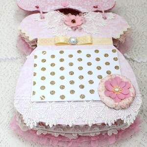 May include: A pink and white baby dress scrapbook with a gold polka dot card, a yellow ribbon, and a pearl embellishment.