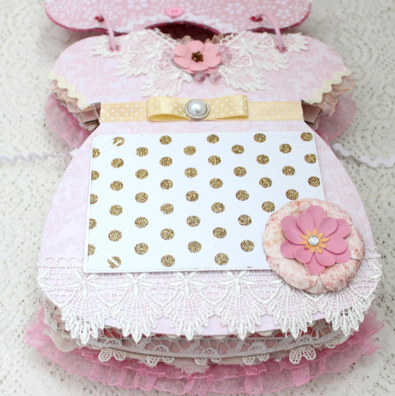 May include: A pink and white baby dress scrapbook with a gold polka dot card, a yellow ribbon, and a pearl embellishment.