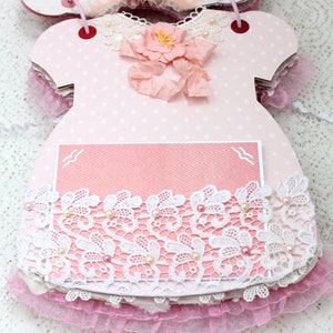 May include: Pink and white baby dress scrapbook with a lace trim and a pink card with a white bird design.