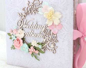 May include: White wedding album with a lace cover and a wooden floral wreath with the words "Wedding album" on it. The wreath is decorated with pink, white, and blue flowers.